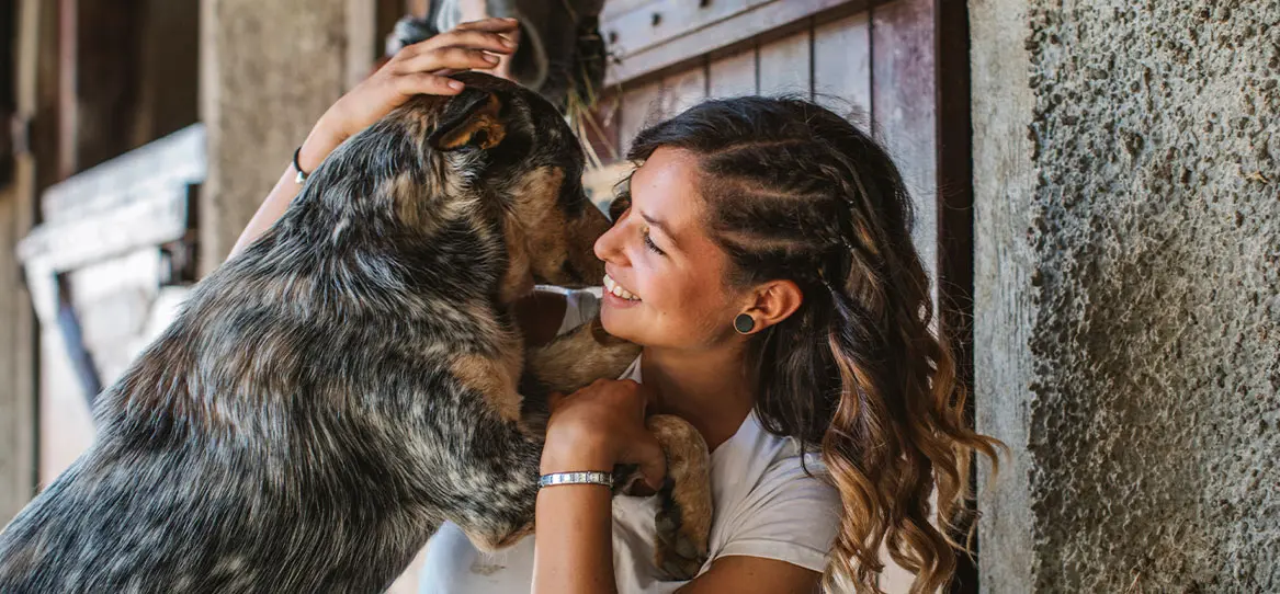 Glückliche jüngere Frau, die mit Ihrem Hund spielt, das Ganze vor einer undeutlich erkennbaren Hauswand