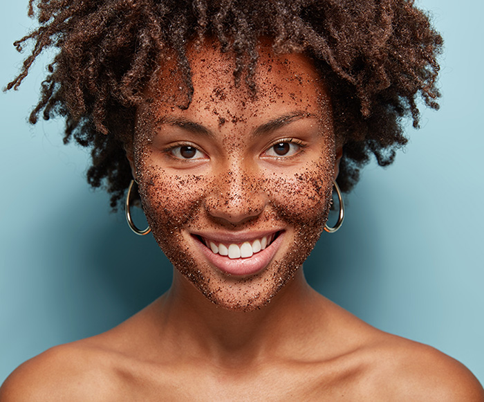 beautiful-mixed-race-female-has-skin-scrub-face-smiles-gently-makes-cosmetic-masks-from-coffee-has-curly-hairstyle-bare-shoulders-isolated-blue-wall_klein Frau mit Kaffee Peeling im Gesicht