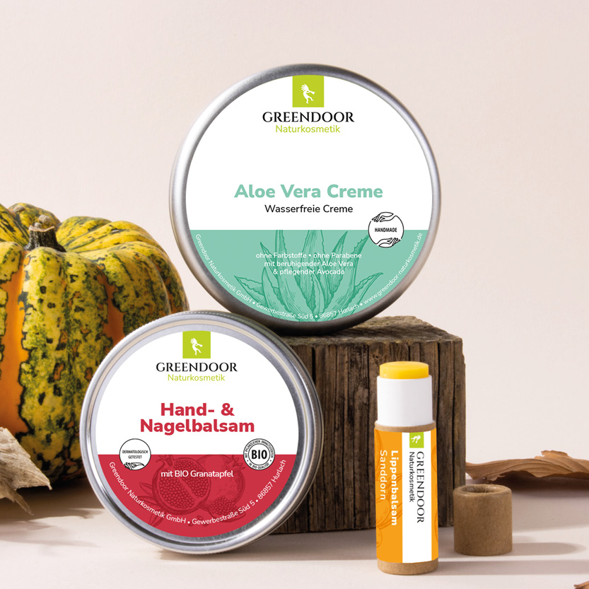 Bildmaterial_Herbst_neu8 Drei Greendoor Produkte vor herbstlichem Hintergrund mit einem Kürbis und Holz. Zu sehen sind Greendoor Handbalsam, Aloe Vera Creme und Lippenbalsam