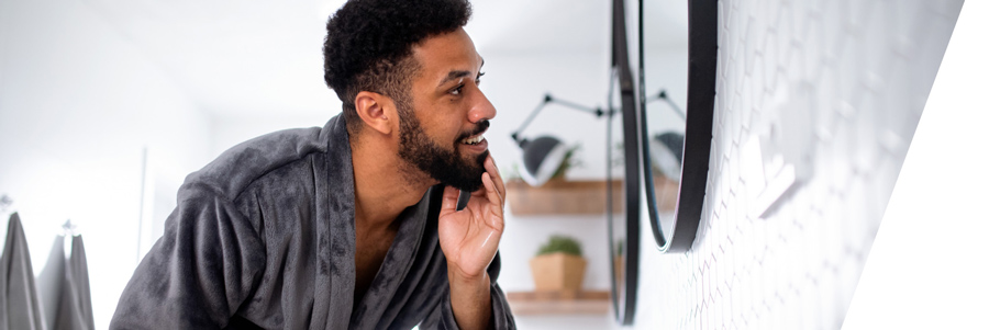 Junger farbiger Mann im grauen Bademantel schaut nach rechts in einen dort hängenden Badezimmerspiegel