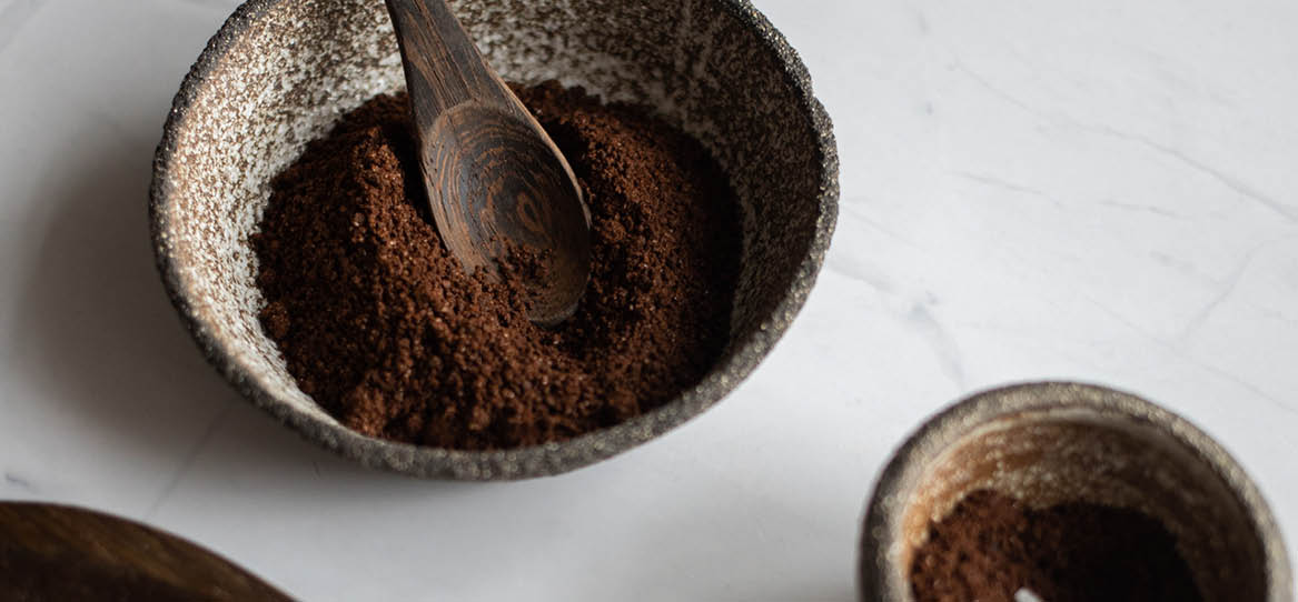 Braunes Pulver, vielleicht gemahlener Kaffee befindet sich in einer kleinen rustikalen Tonschüssel zusammen mit einem rustikalen einfachen Holzlöffel