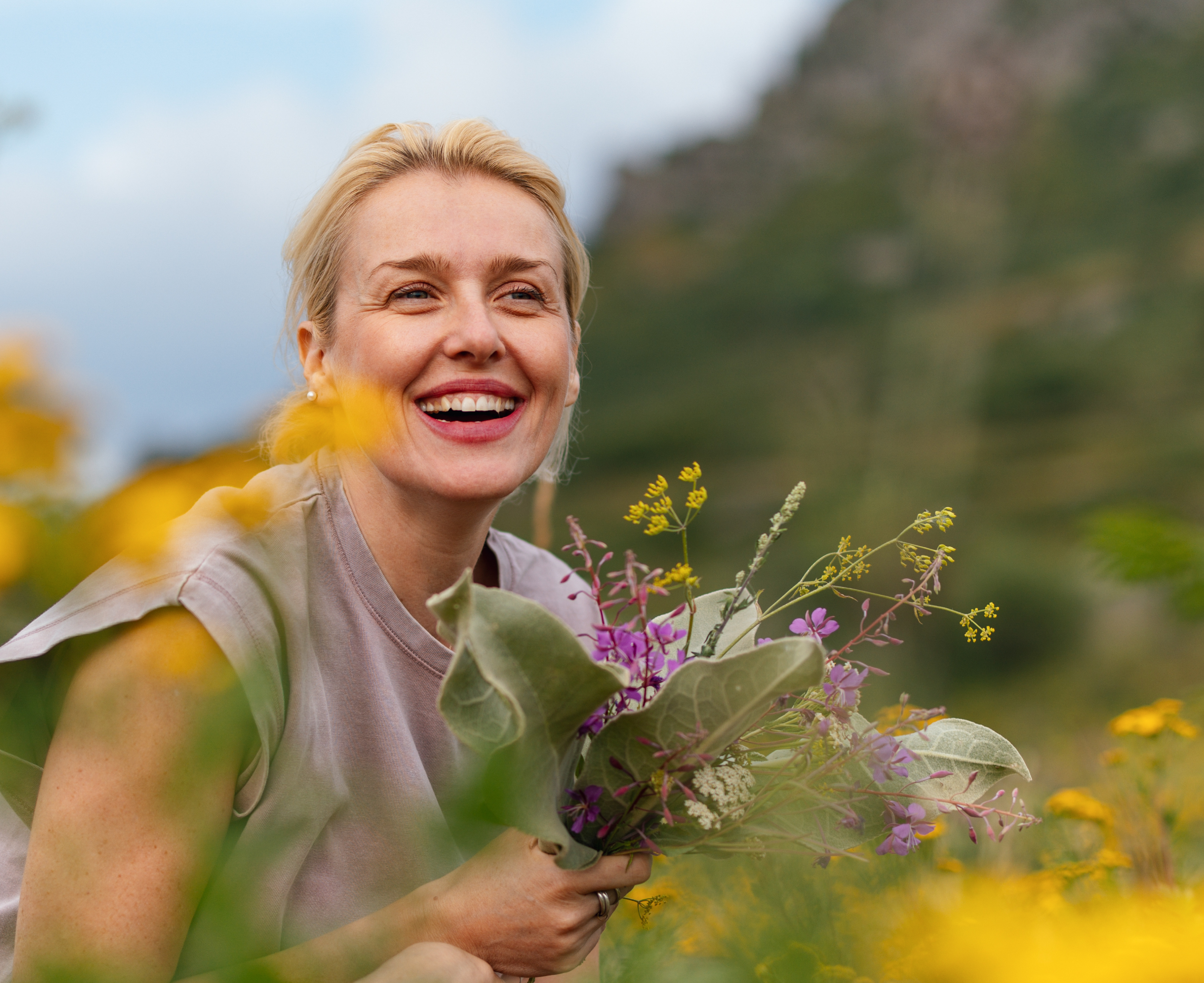 Frau mittleren Alters lacht in einer Wiese stehend und hat einen Wildblumenstrauß