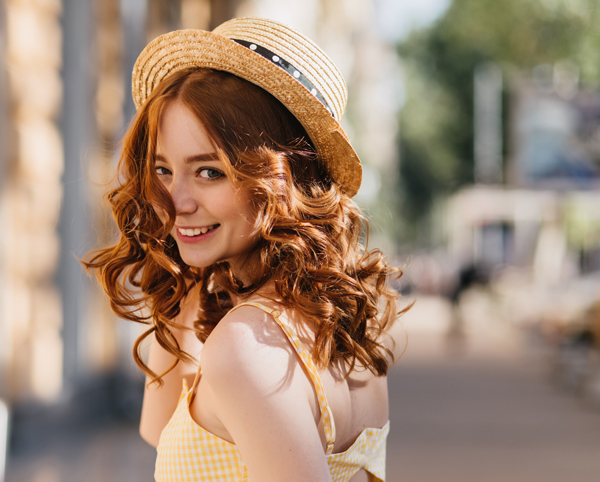 enchanting-girl-with-dark-curly-hair-urban-street junge lächelnde Frau mit Sommerhut und langen lockigen roten Haaren vor unscharfem Hintergrund