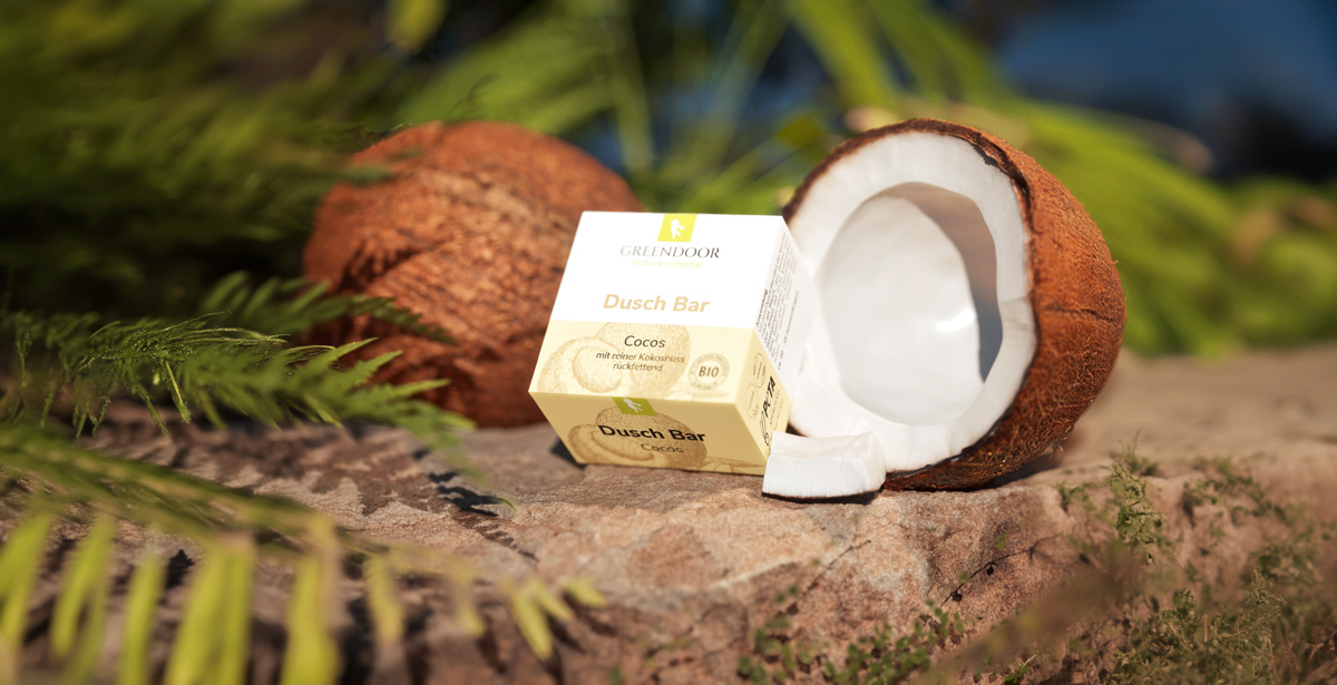 Duschbar_Cocos_3D_Visualisierung Kokosnüsse, davon eine geöffnet zusammen mit dem Greendoor Dusch Bar Cocos platziert auf einem Fels, der von Grün umgeben ist