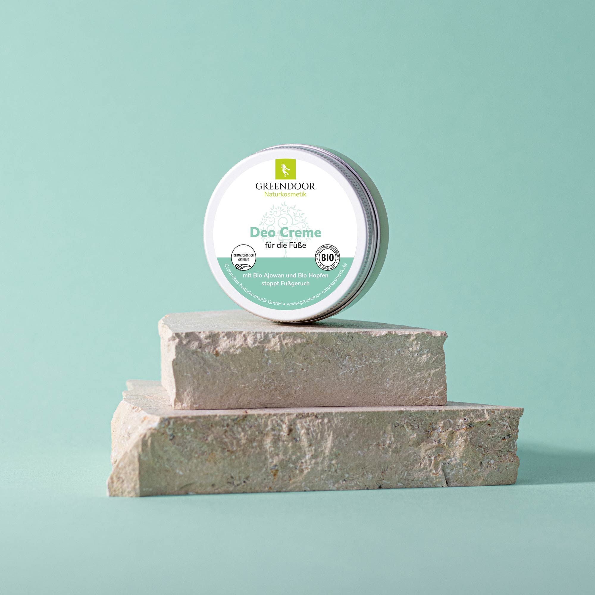 Eine Dose Greendoor Deo Creme für die Füße steht auf einem Steinpodest vor einem mintgrünen Hintergrund