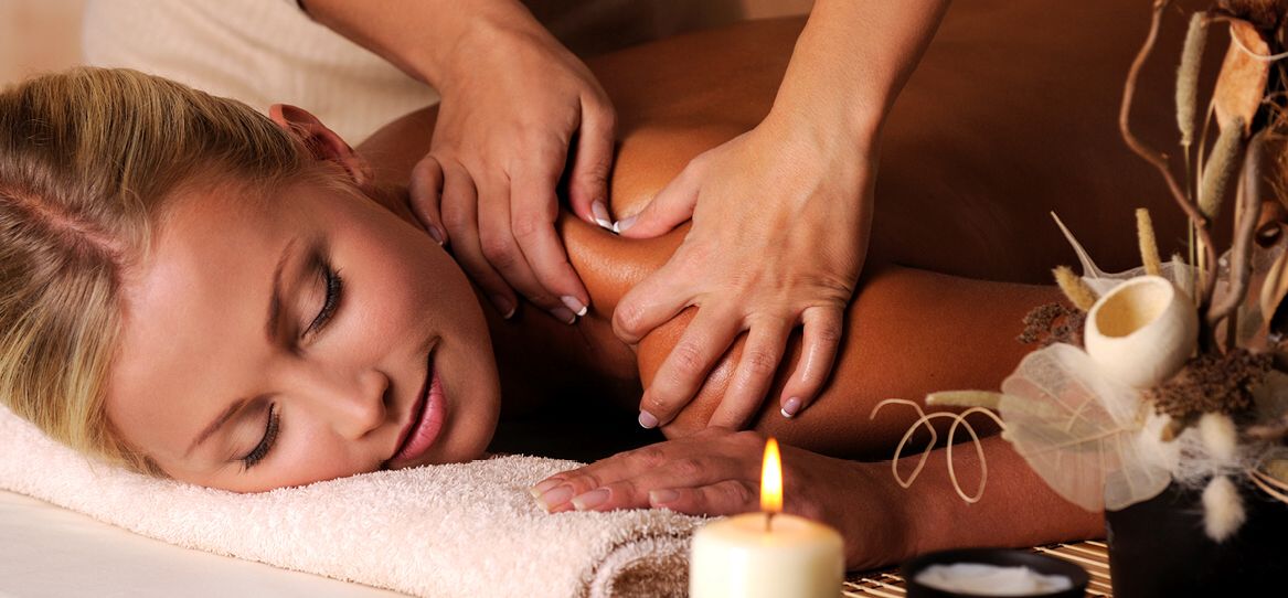 Junge blonde liegende Frau mit geschlossenen Augen und sanftem Lächeln wird gerade von einer Masseurin massiert