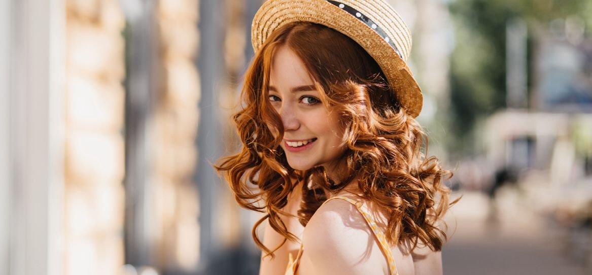 Junge Frau mit roten langen sehr lockigen Haaren und einem Sommerhut lächelt fröhlich über die Schulter schauend in die Kamera